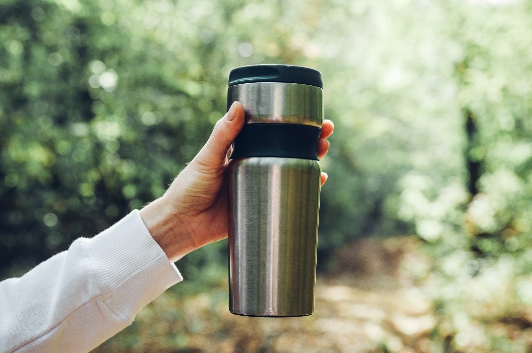 Unrecognizable woman traveler holding thermos mug on background of green forest. Travel, hiking, sport, healthy lifestyle concept